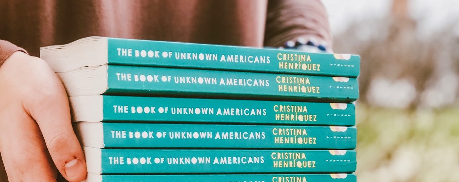 Story Image - Student holding stack of books titled The Book of Unknown Americans
