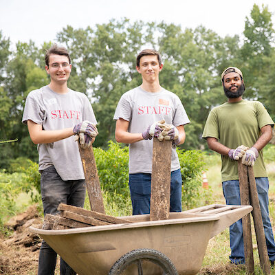 Three Witt Fellows hold wood and stand behind a wheelbarrow.