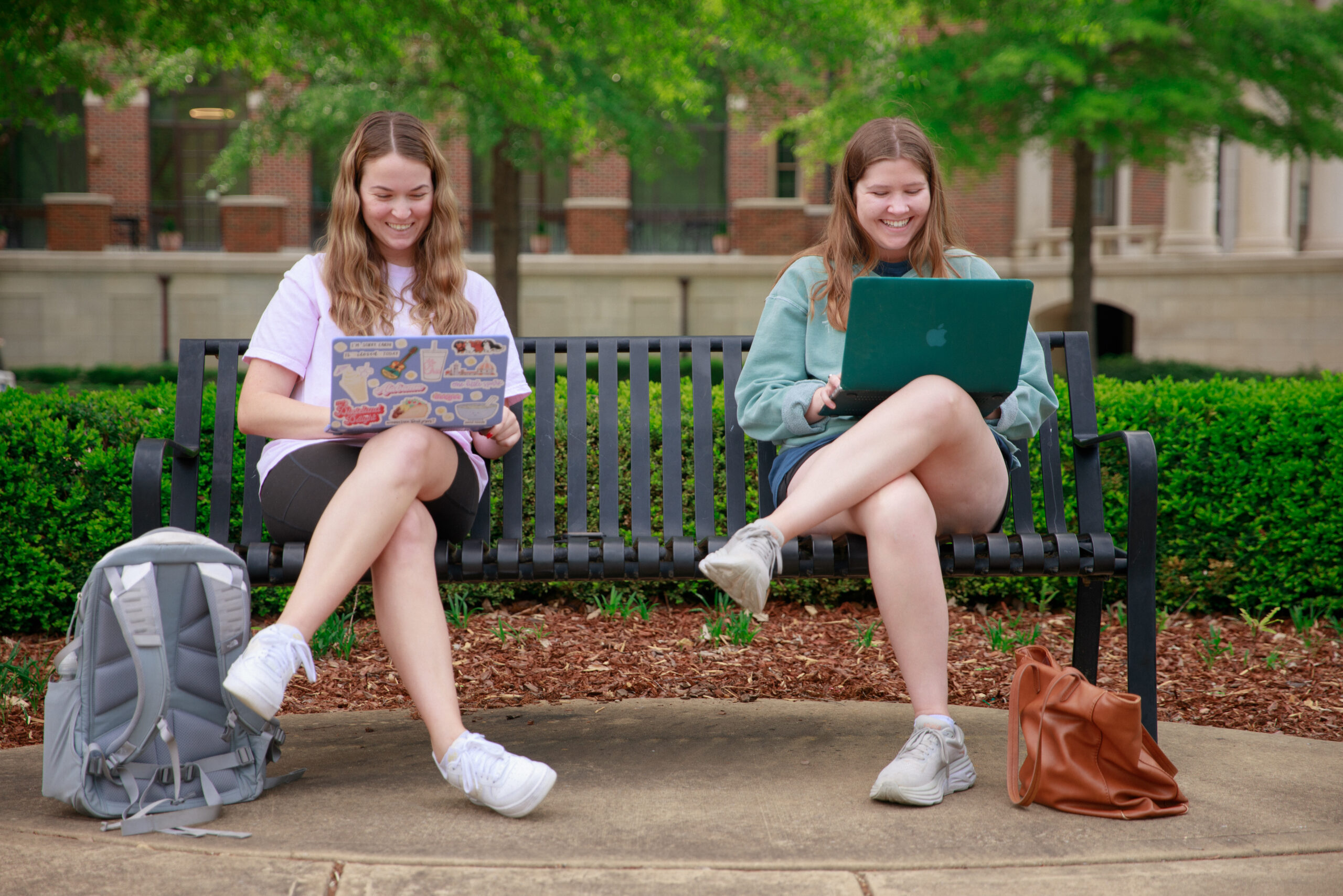 Two students sit on a bench and smile while on laptops.