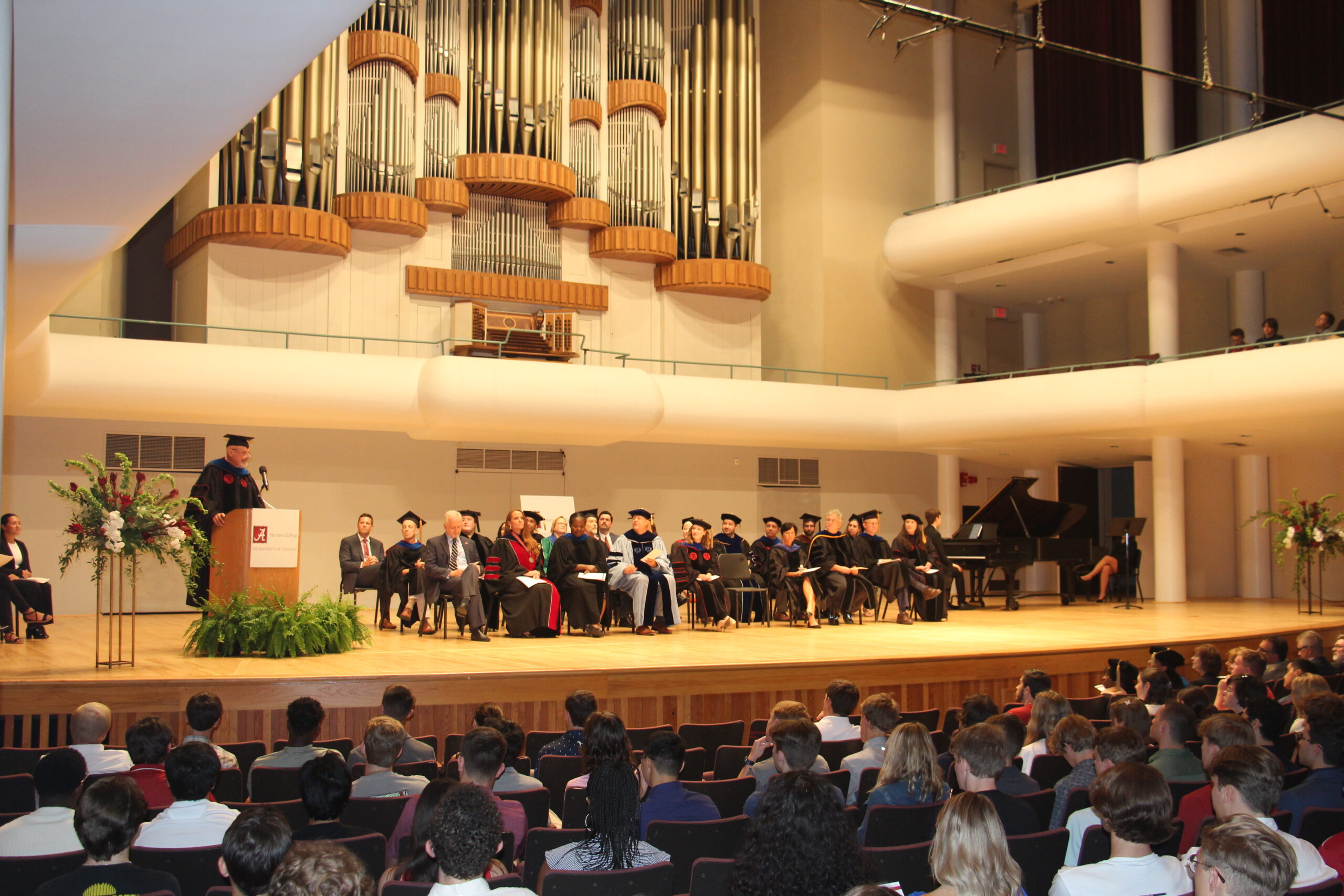 Honors College faculty and staff in regalia on stage while Dr. John Latta speaks at a podium.