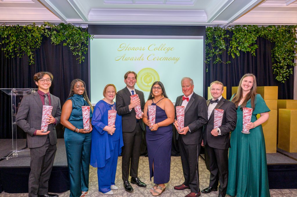 A photo of all 2025 award recipients, smiling with their awards, crimson and white glass vases.